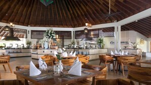 Een restaurant met houten tafels en stoelen, een hoog houten plafond en een keuken in de achtergrond.