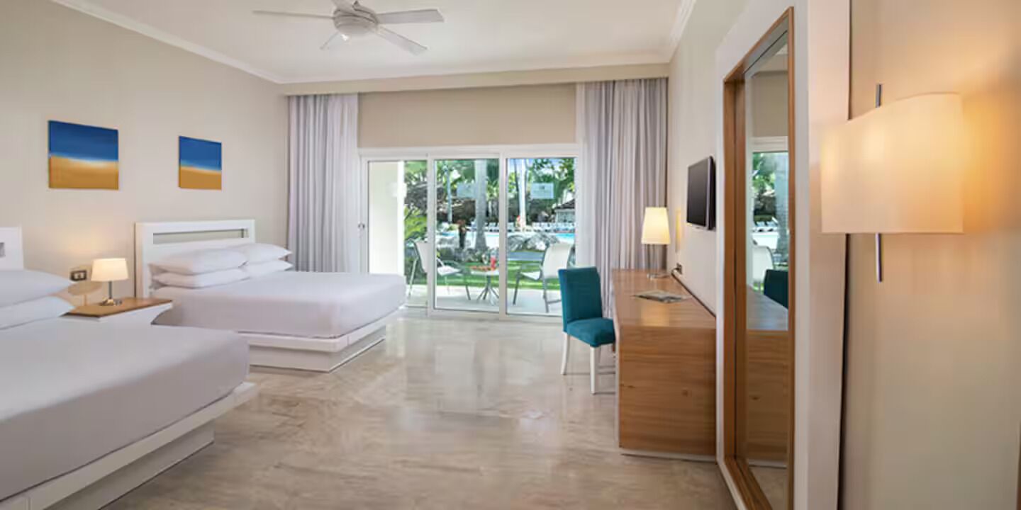 Een hotelkamer met twee bedden, een bureau, een stoel, een plafondventilator en uitzicht op de omgeving.