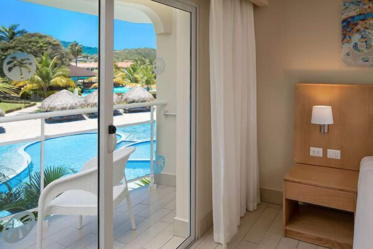 Ein Hotelzimmer mit Bett, Nachttisch mit Lampe und Glastür, die zu einem Balkon mit Blick auf einen Pool führt.