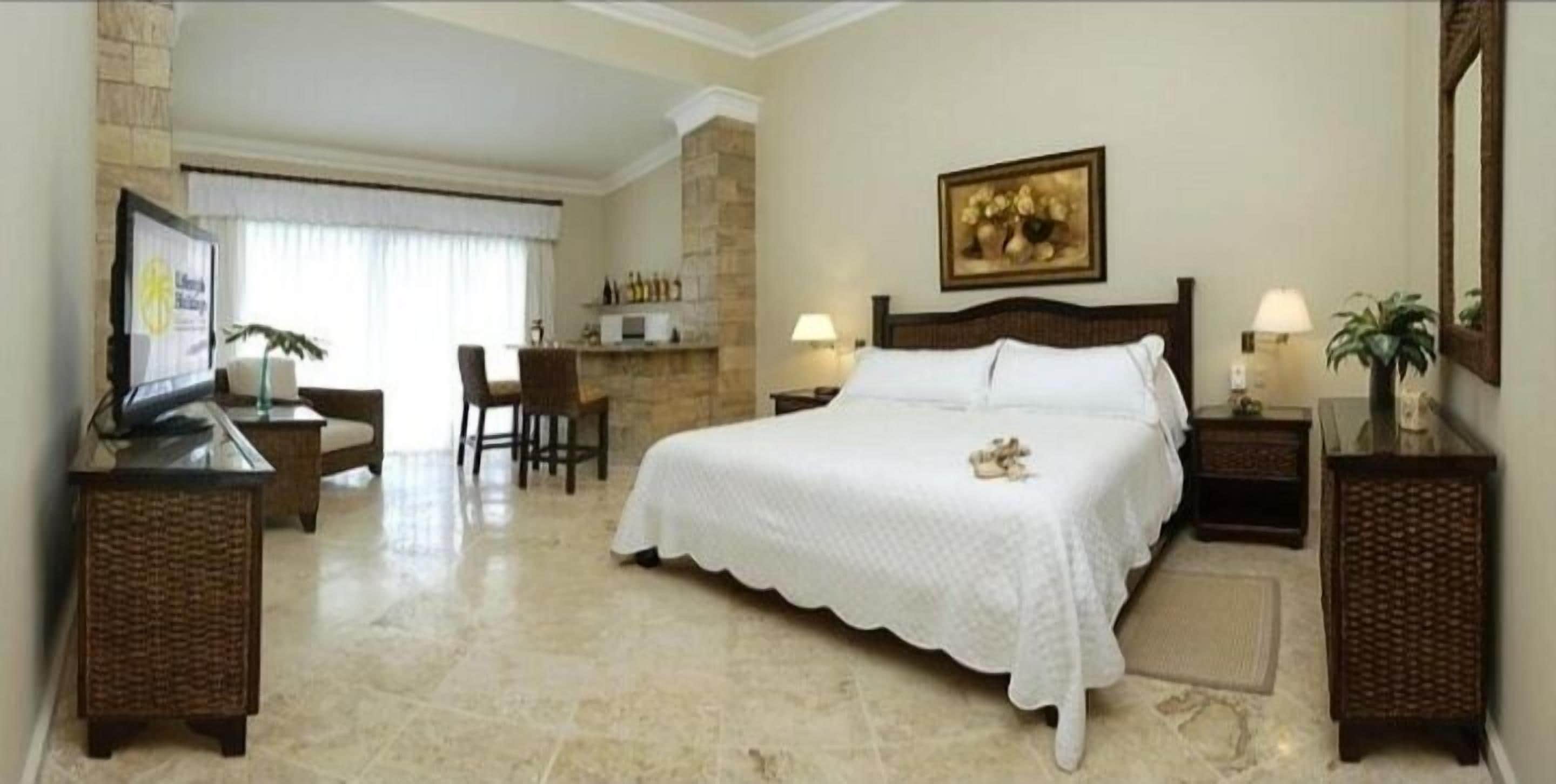 Habitación de hotel con una cama grande, un escritorio con ordenador, una silla, una mesita con botellas y un televisor montado en la pared.
