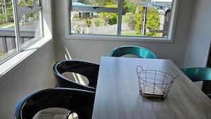 A dining area with a table and chairs, large windows with a view of a residential area, and a wire basket on the table. - Dog friendly Northland getaway - Hihi Beach (Hihi)