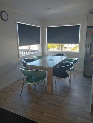 A dining area with a table and chairs, large windows with blinds, a clock on the wall, and a refrigerator. - Dog friendly Northland getaway - Hihi Beach (Hihi)