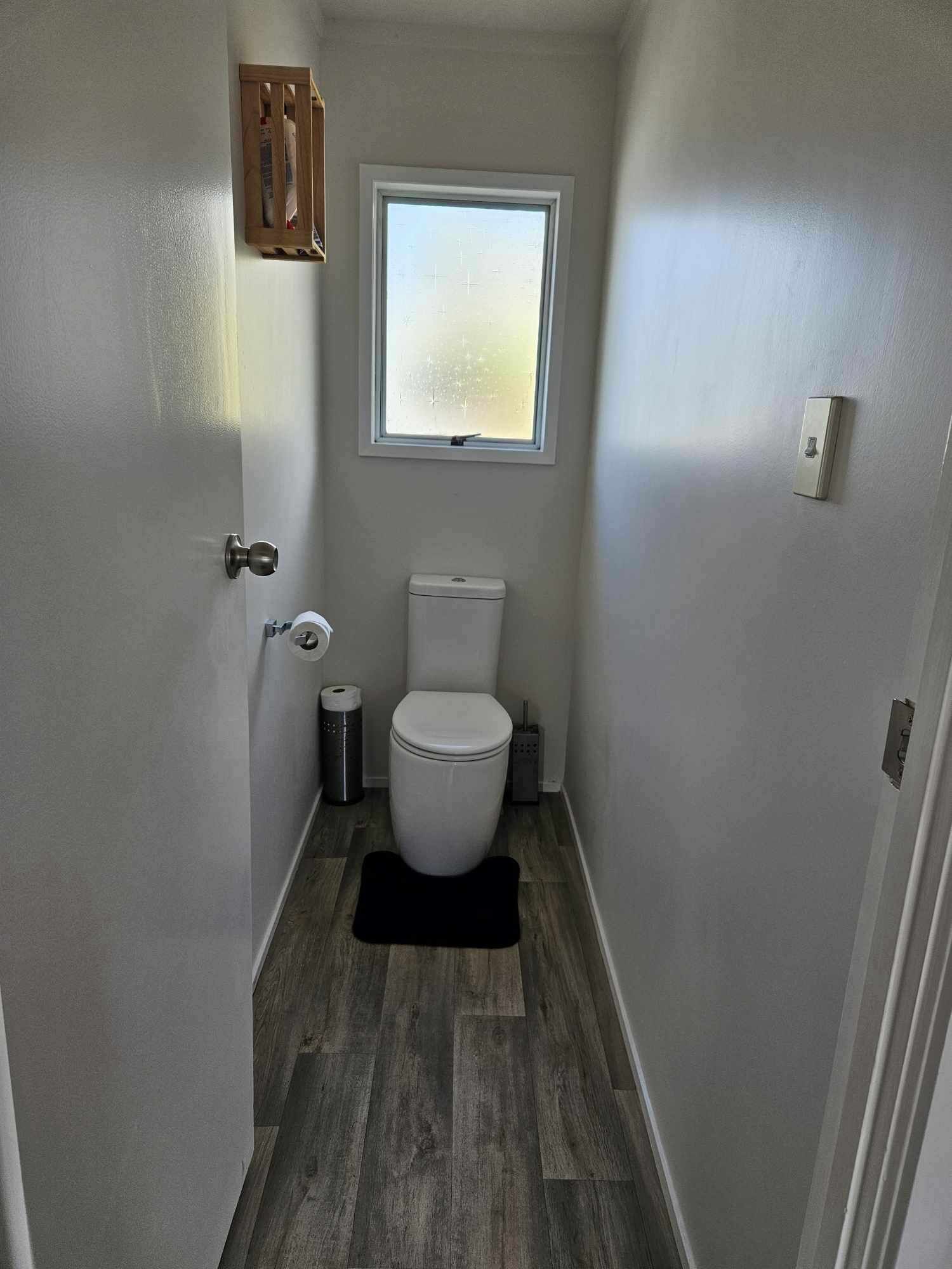 A narrow bathroom with a toilet, a window, and a wooden cabinet.