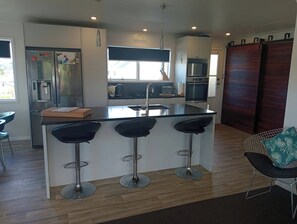 A modern kitchen with a central island, bar stools, and a dining area with a chair and table. - Dog friendly Northland getaway - Hihi Beach (Hihi)