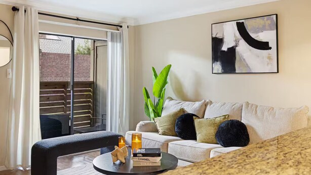 Un salon moderne avec un canapé, une table basse agrémentée de livres et de bougies, une grande plante et une porte-fenêtre coulissante donnant sur un balcon.