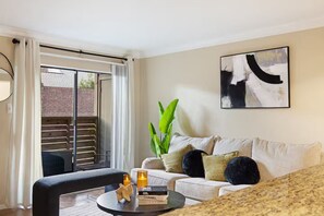 A modern living room with a sofa, a coffee table with books and candles, a large plant, and a sliding glass door leading to a balcony. - 2BR•2BA Steps away from Med Center & NRG Pool &Parking (Houston)