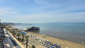 Eine Uferpromenade mit mehreren Reihen Sonnenliegen und Sonnenschirmen, ein Steg, der ins Meer führt, und ein klarer Himmel.