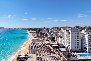Un paysage urbain côtier avec un hôtel en bord de mer, doté de rangées de chaises longues et d’une piscine.