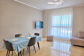 A modern dining area with a table set for two, a flat-screen TV mounted on the wall, and a sofa in the background. - Sea View Apartment in Durres Beach 100, Durres, Albania (Durres)