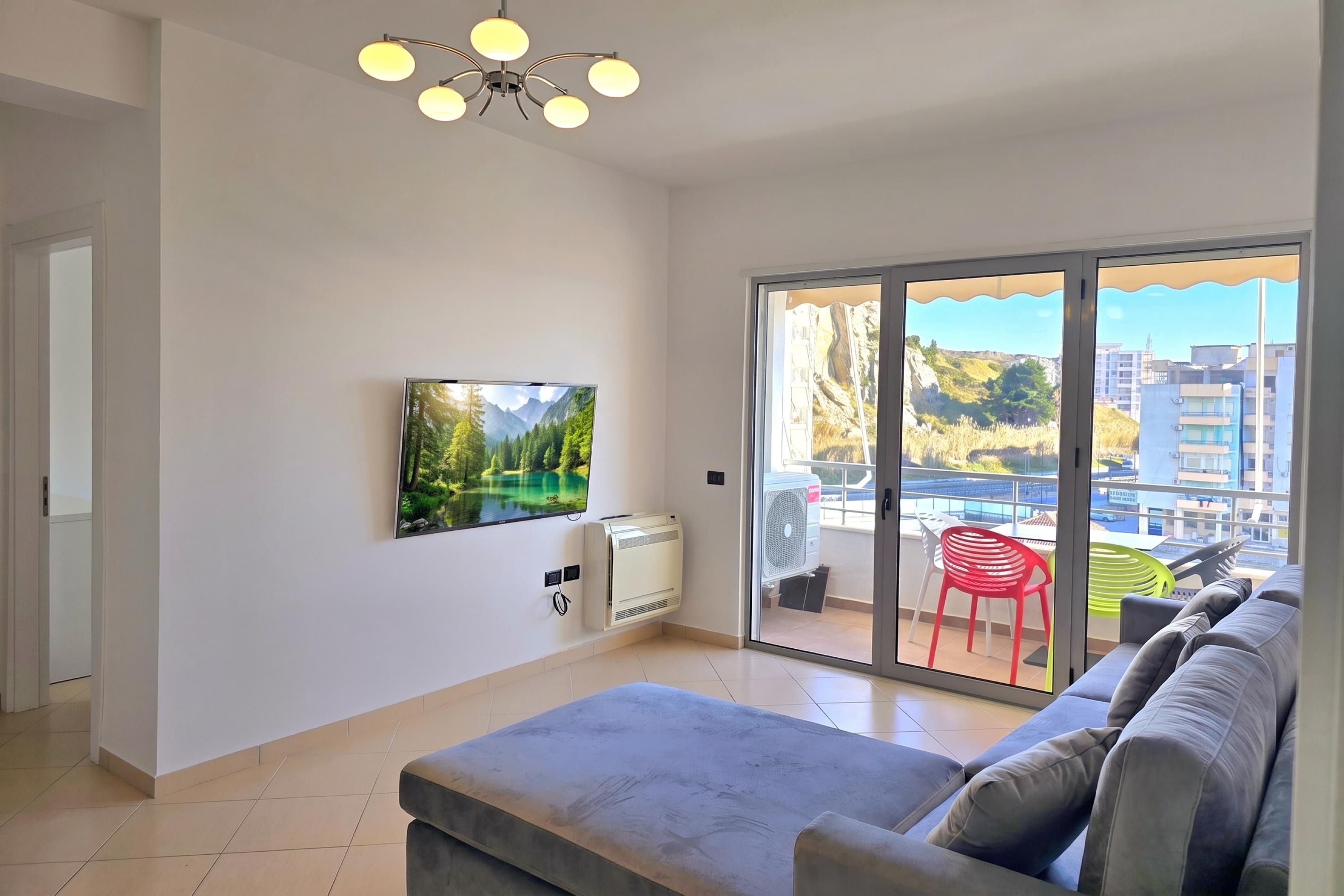 A modern living room with a flat-screen TV mounted on the wall, a grey sofa, and a sliding glass door leading to an outdoor area with a view of buildings and a red chair.