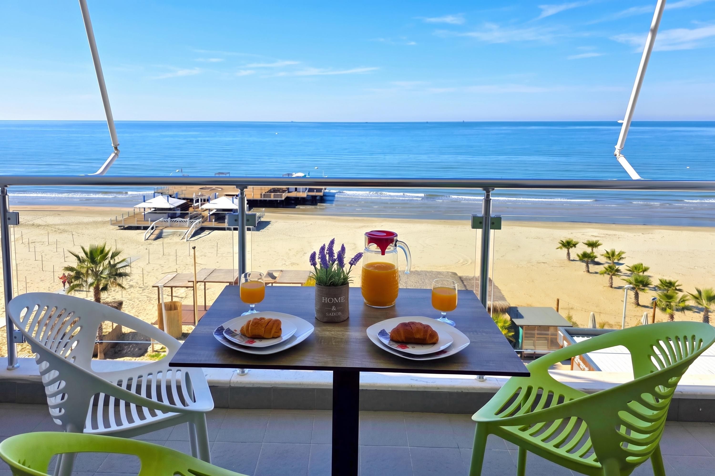 A balcony with a table set for breakfast, overlooking a beach with palm trees and a clear blue sky.
