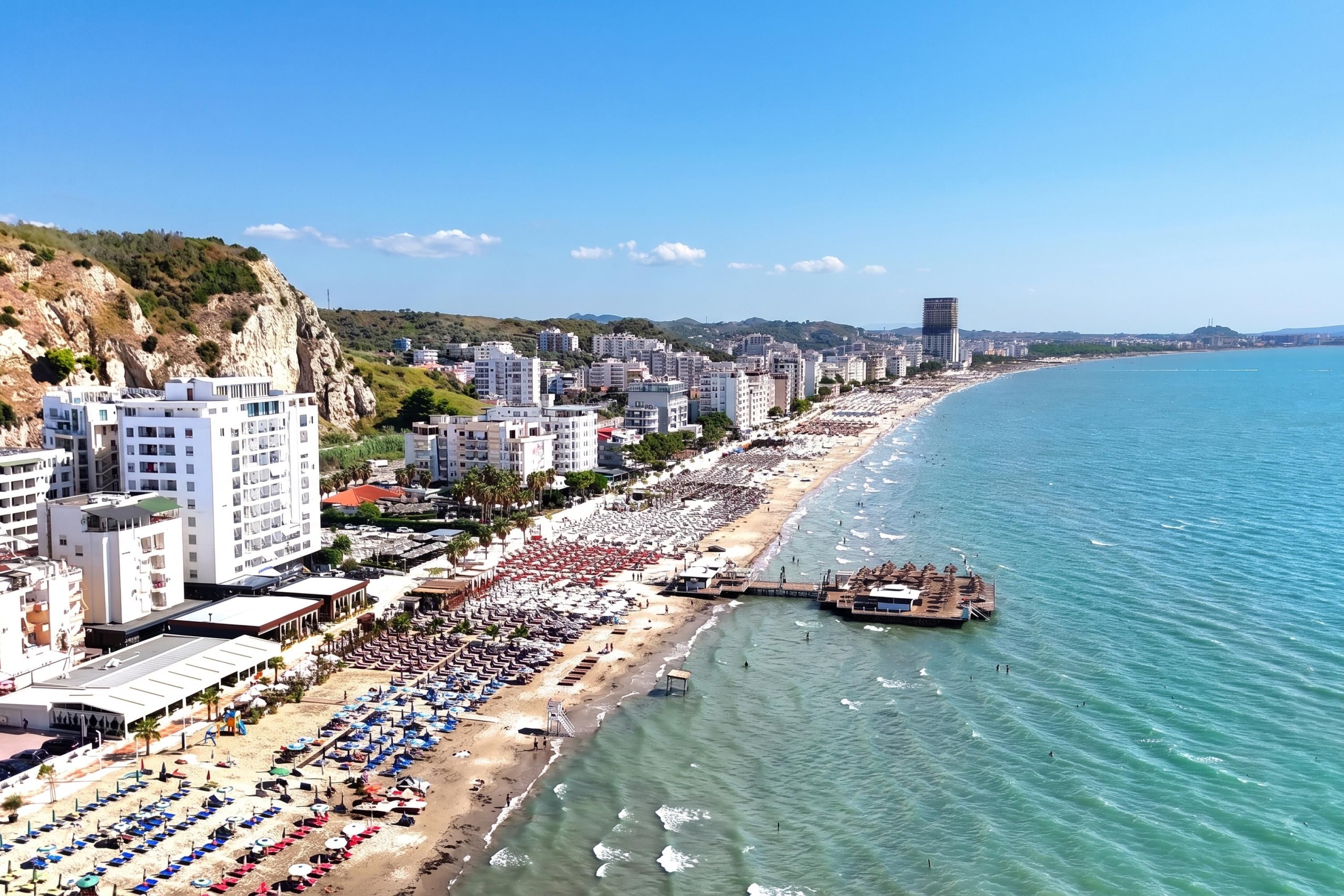 A coastal cityscape with a long beachfront lined with rows of sunbeds and umbrellas, a pier extending into the sea, and a backdrop of modern buildings and hills.