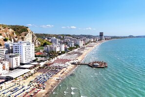 Un paisaje urbano costero con una extensa playa bordeada por filas de tumbonas y sombrillas, un muelle que se adentra en el mar, y un telón de fondo de edificios modernos y colinas.