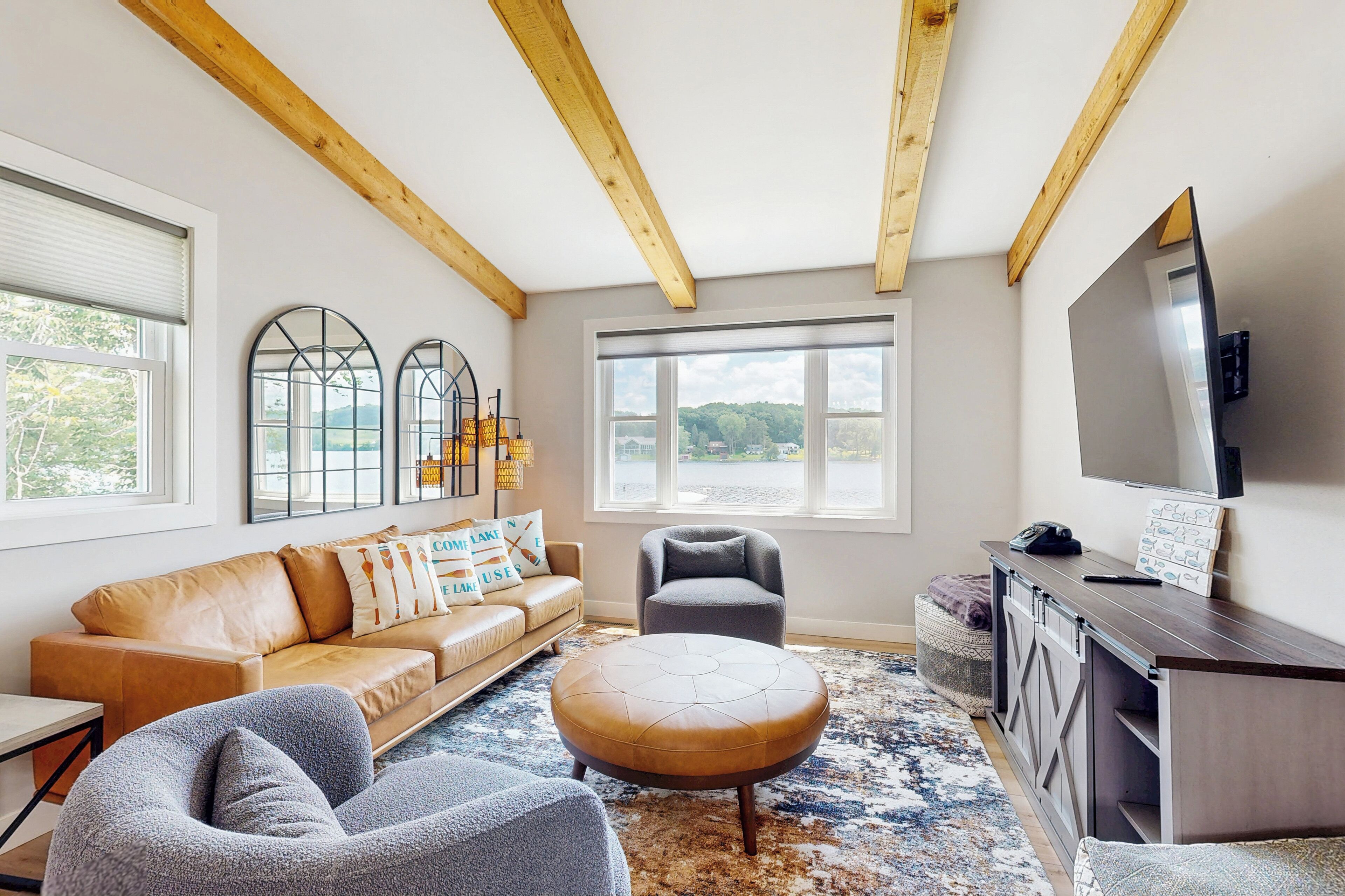 Un salon comprenant un canapé, un pouf, un fauteuil et un téléviseur posé sur un meuble en bois. De grandes fenêtres offrent une vue sur l’extérieur.