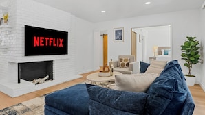 Een moderne woonkamer met een wit bakstenen haardvuur, een flatscreen-tv waarop 'NETFLIX' wordt weergegeven, een blauwe bank, een witte fauteuil en een bijzettafeltje.
