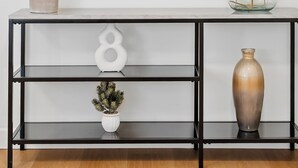 Een moderne console tafel met een spiegel erboven, aan weerszijden een potplant en een vaas met gedroogde bloemen.