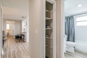 Een gang die leidt naar een badkamer met toilet en bad, een klein zithoekje met een tafel, en een kast met planken.