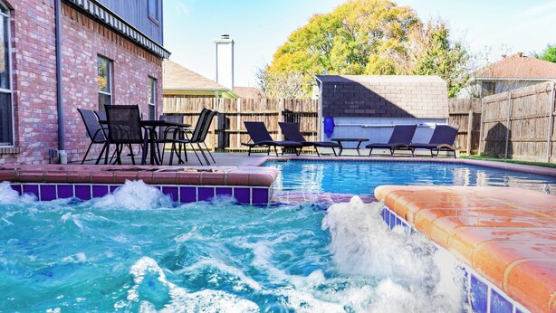 A swimming pool with lounge chairs and a hot tub, surrounded by a wooden fence and a brick building. - Sleeps 12 - Pool, Hot Tub & Game Room (Grand Prairie)
