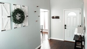 A hallway with a light blue wall, wooden floor, and a white door with a semi-circular window. There are three white barn-style shutters on the wall and a wreath above them. - Sleeps 12 - Pool, Hot Tub & Game Room (Grand Prairie)