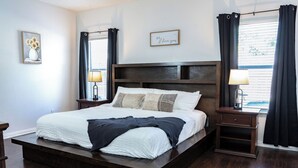 A bedroom with a large bed, dark wood headboard, two bedside tables with lamps, a small nightstand, and a window with curtains. - Sleeps 12 - Pool, Hot Tub & Game Room (Grand Prairie)