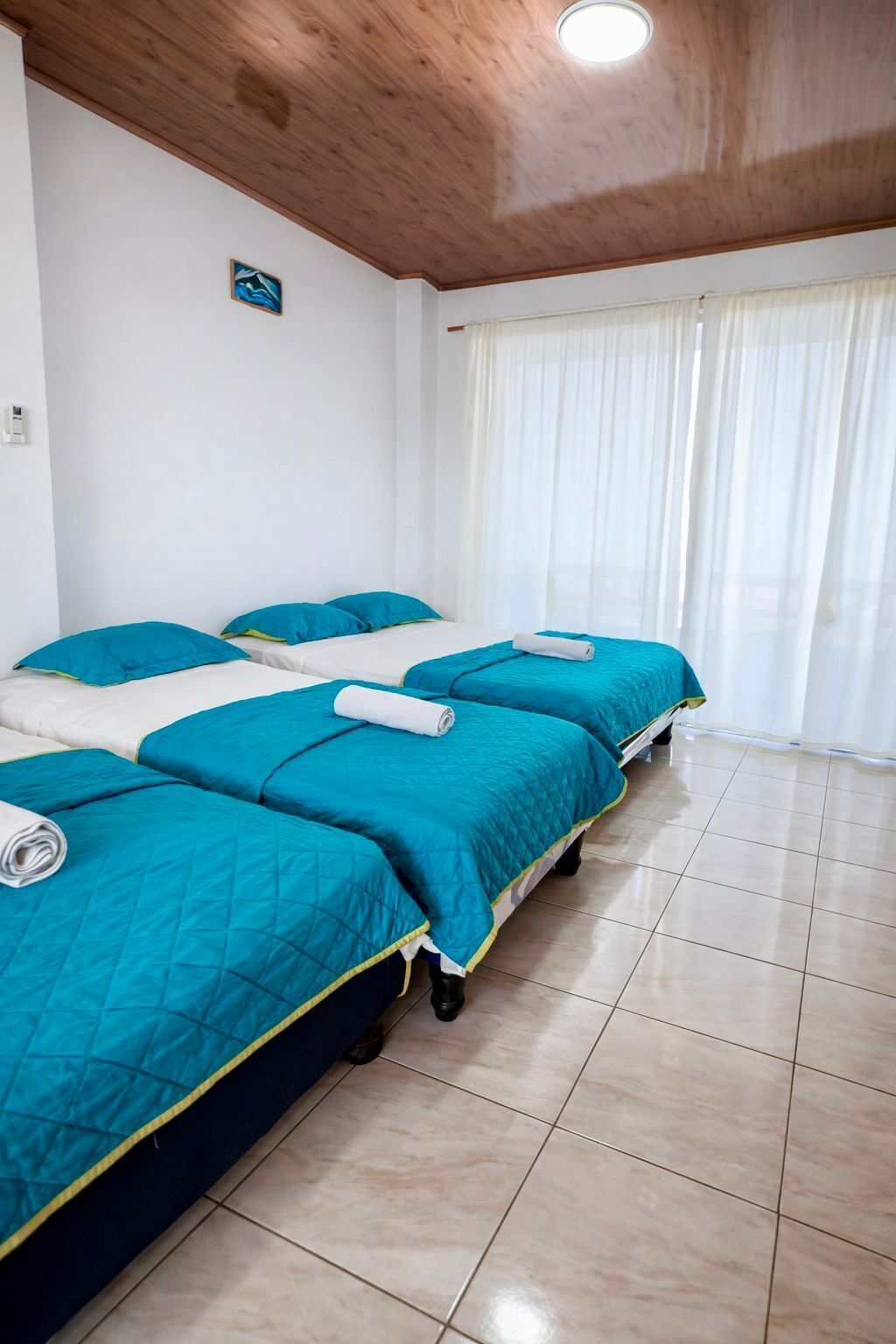 A hotel room with two beds, each with teal bedding and white pillows, a wooden ceiling, and a large window with sheer curtains.