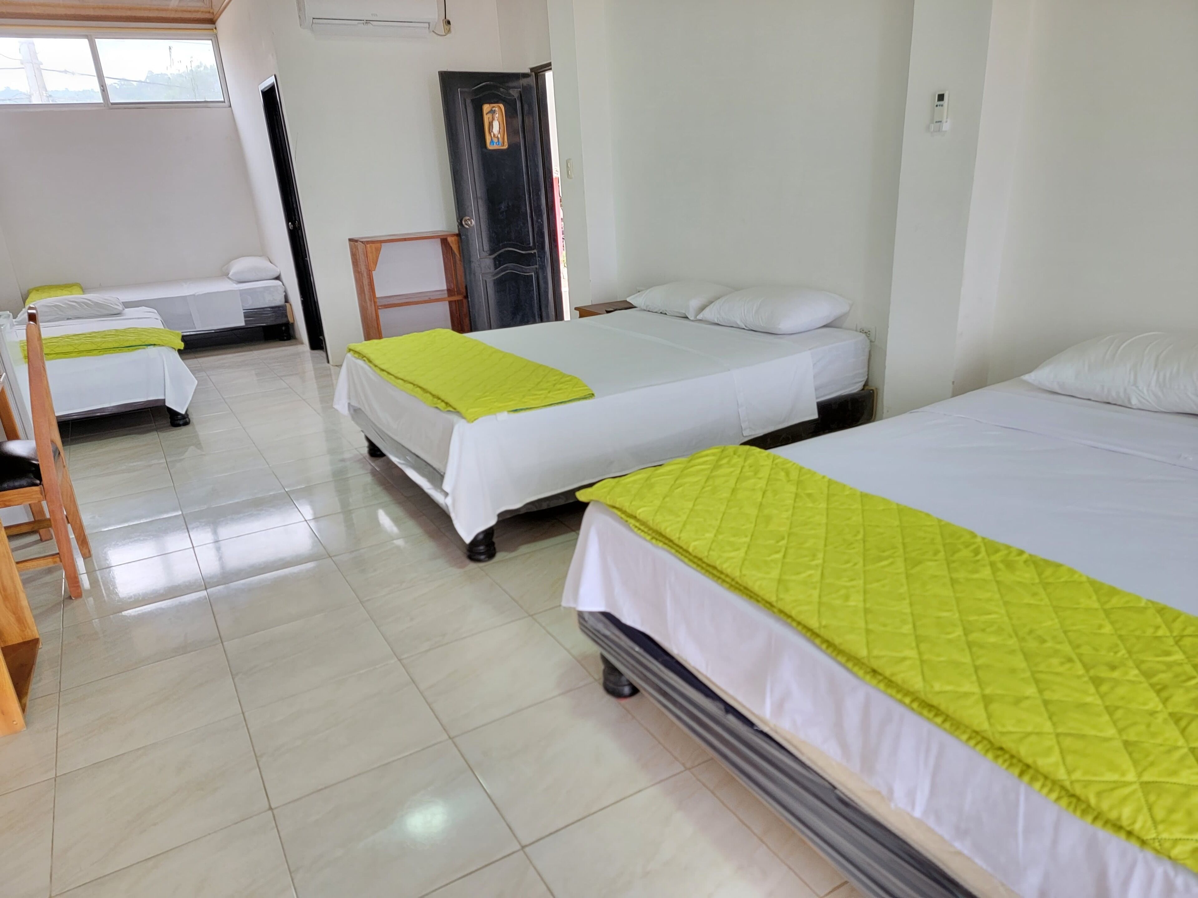Habitación de hotel con tres camas individuales, cada una con colchón blanco y colcha verde. Hay una silla de madera y una mesita en la habitación.