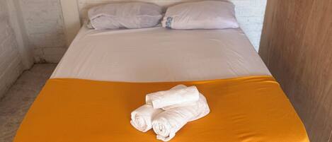 Una cama con colchón y almohadas blancas, una manta amarilla y dos toallas enrolladas sobre la cama.
