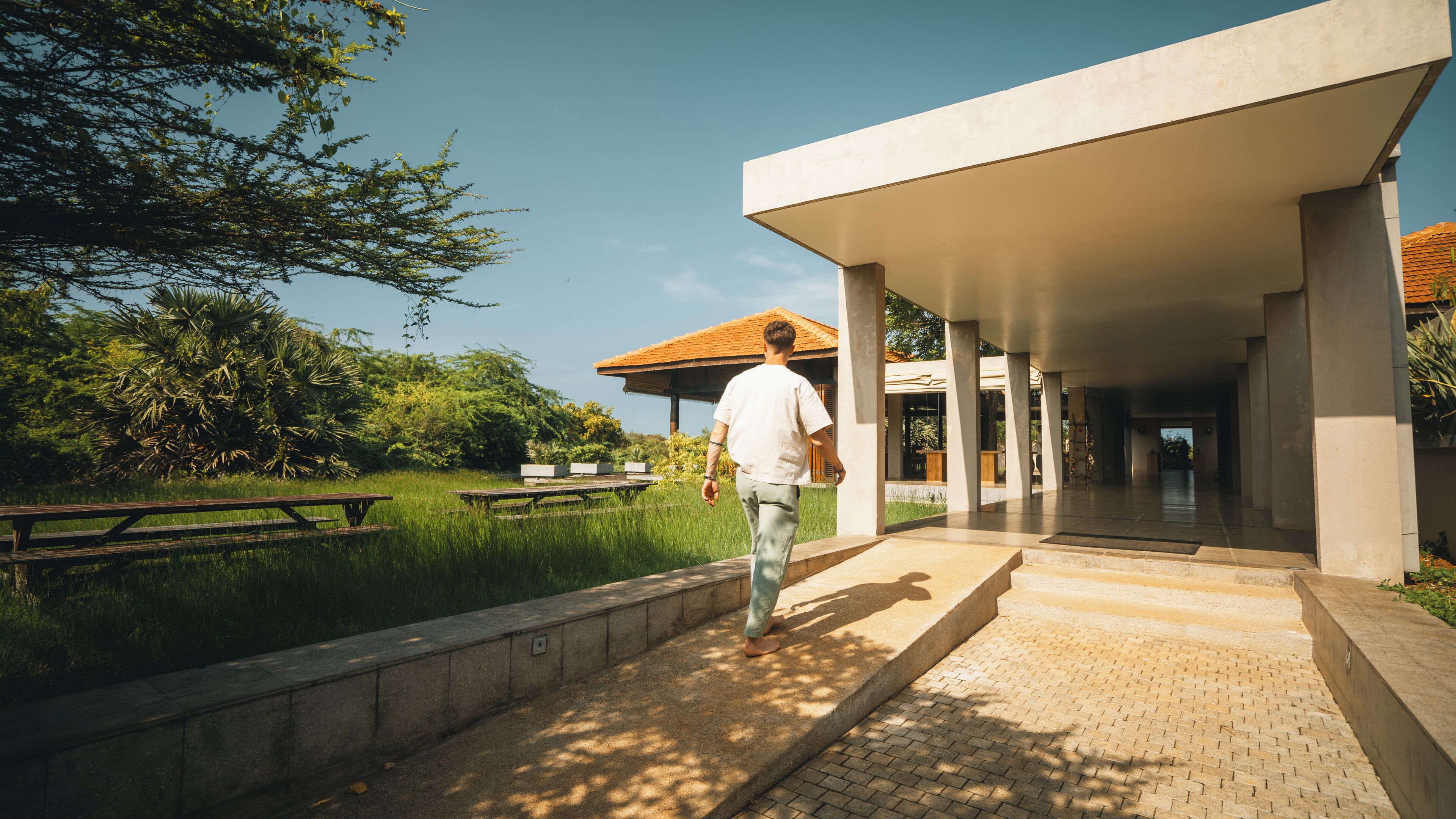 A man walking towards a covered outdoor area with picnic tables and a building with a tiled roof.