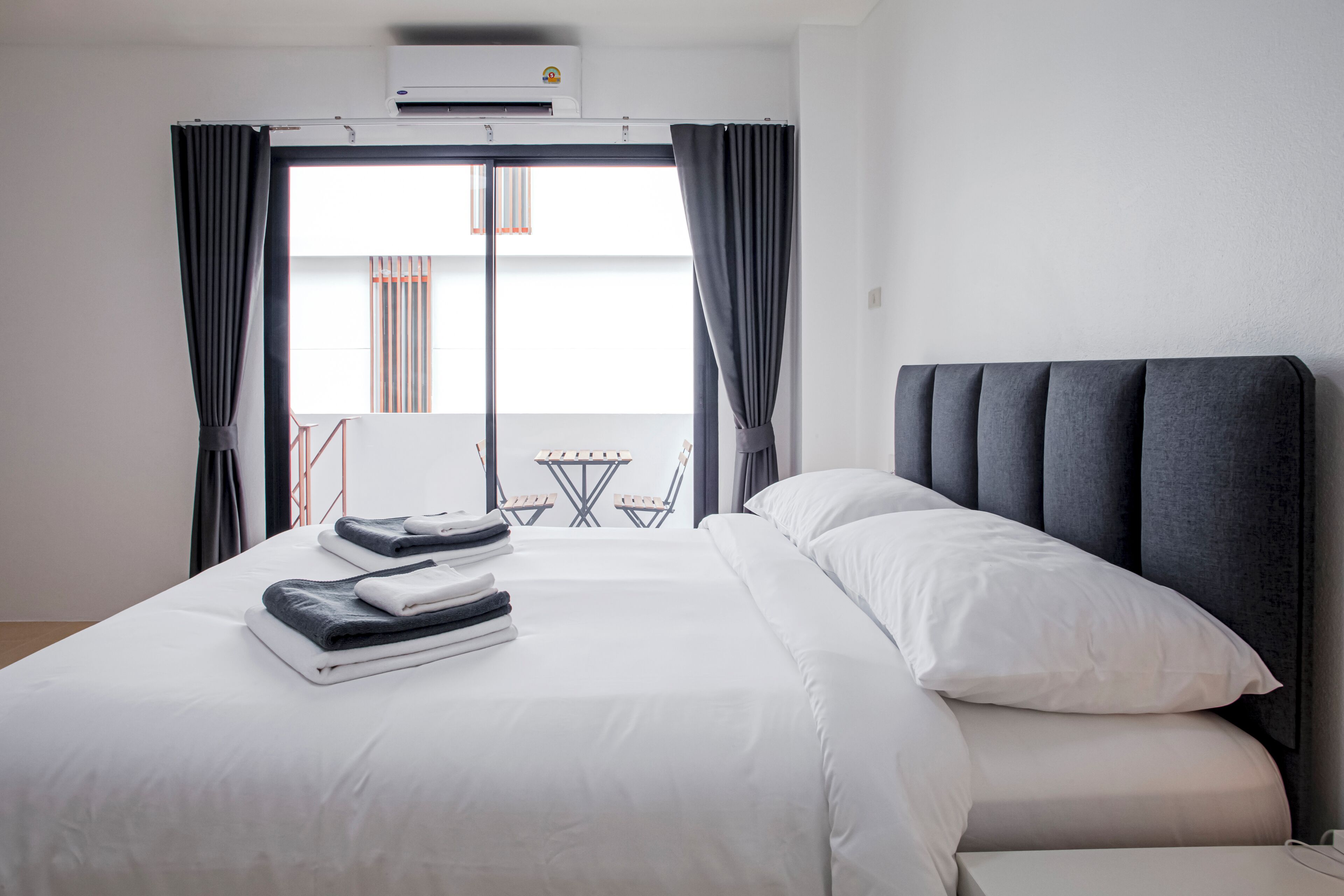 Una habitación de hotel con una cama, un ventanal con cortinas, una pequeña mesa y sillas afuera de la ventana y un equipo de aire acondicionado en la pared.