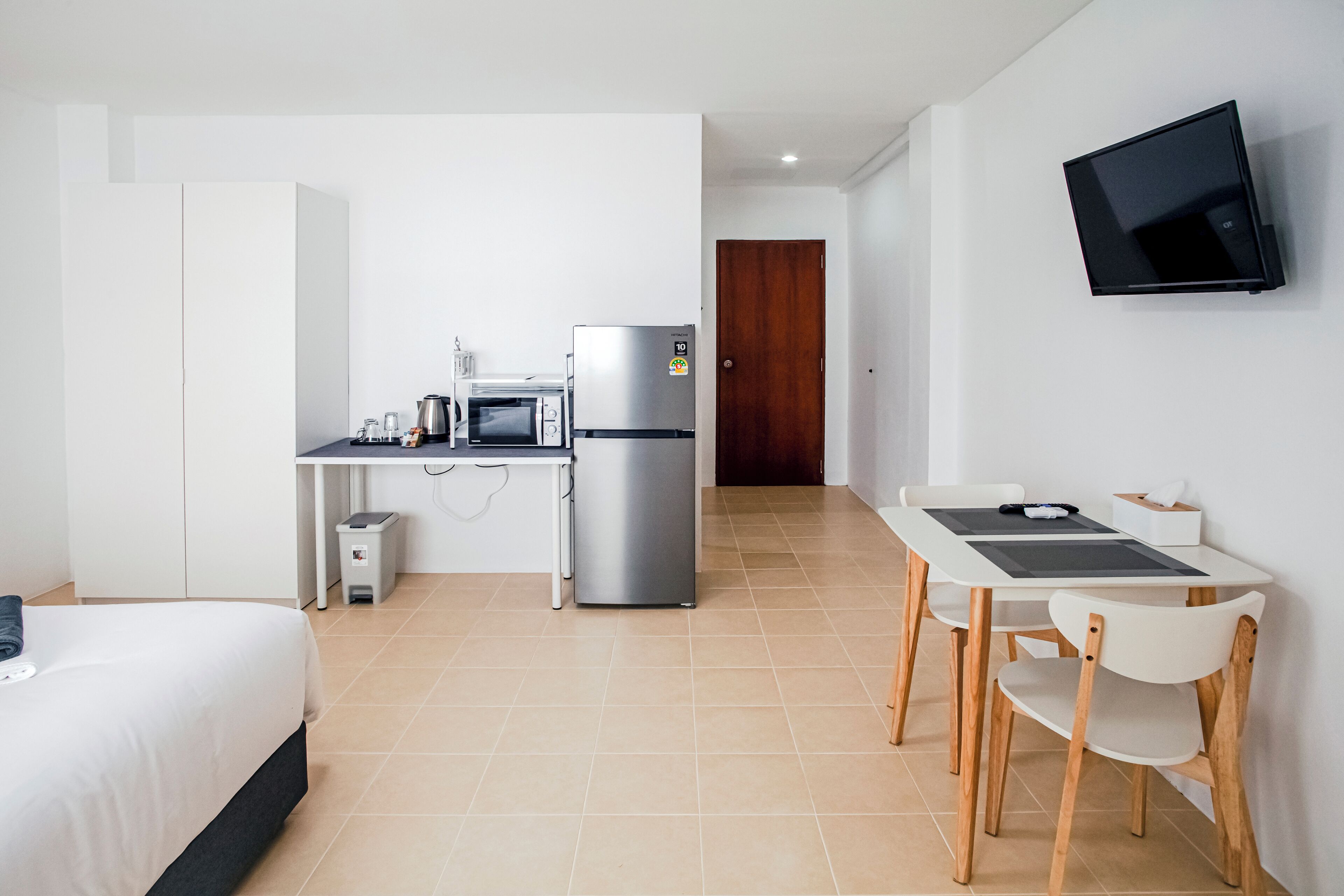 A hotel room with a bed, a kitchenette with a fridge, microwave, and coffee maker, a dining area with a table and chairs, and a TV mounted on the wall.
