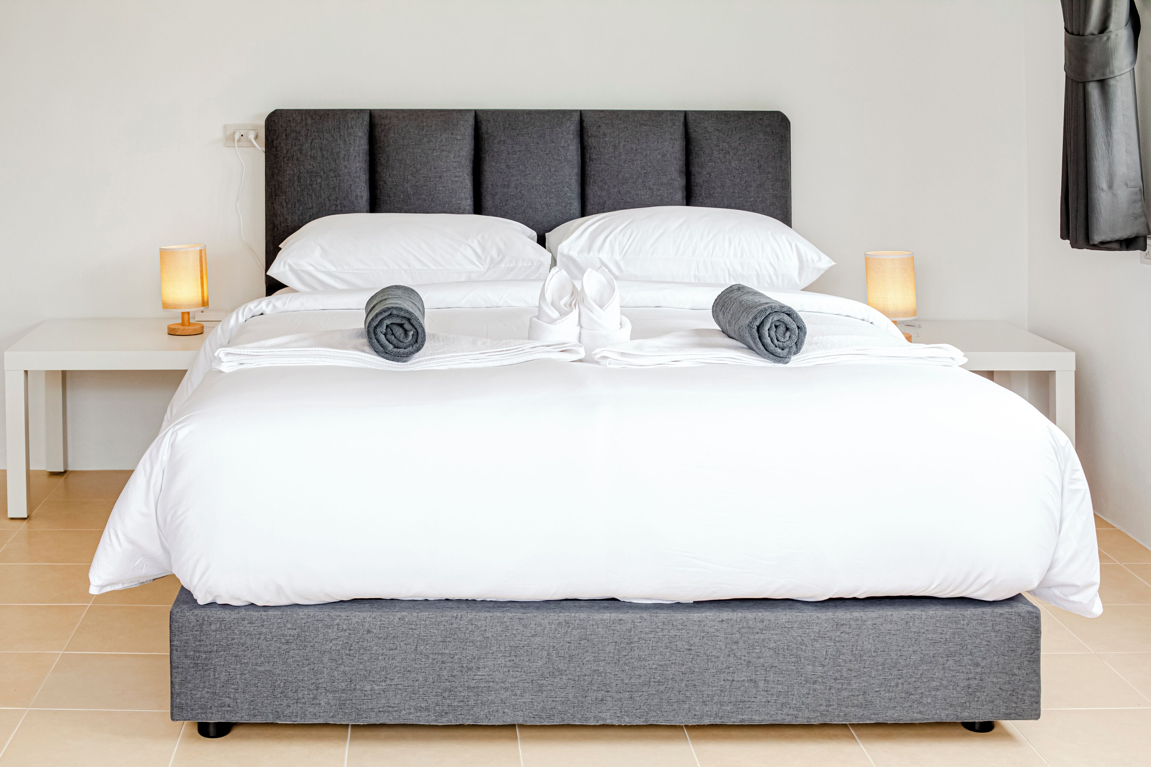 A neatly made bed with white and grey bedding, two rolled towels, and two bedside lamps on white nightstands.