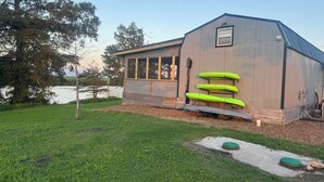 A building with a large window, a kayak rack, and a grassy area with a small pond. - Cozy, lakeside tiny home in Arkansas farm country (Parkin)