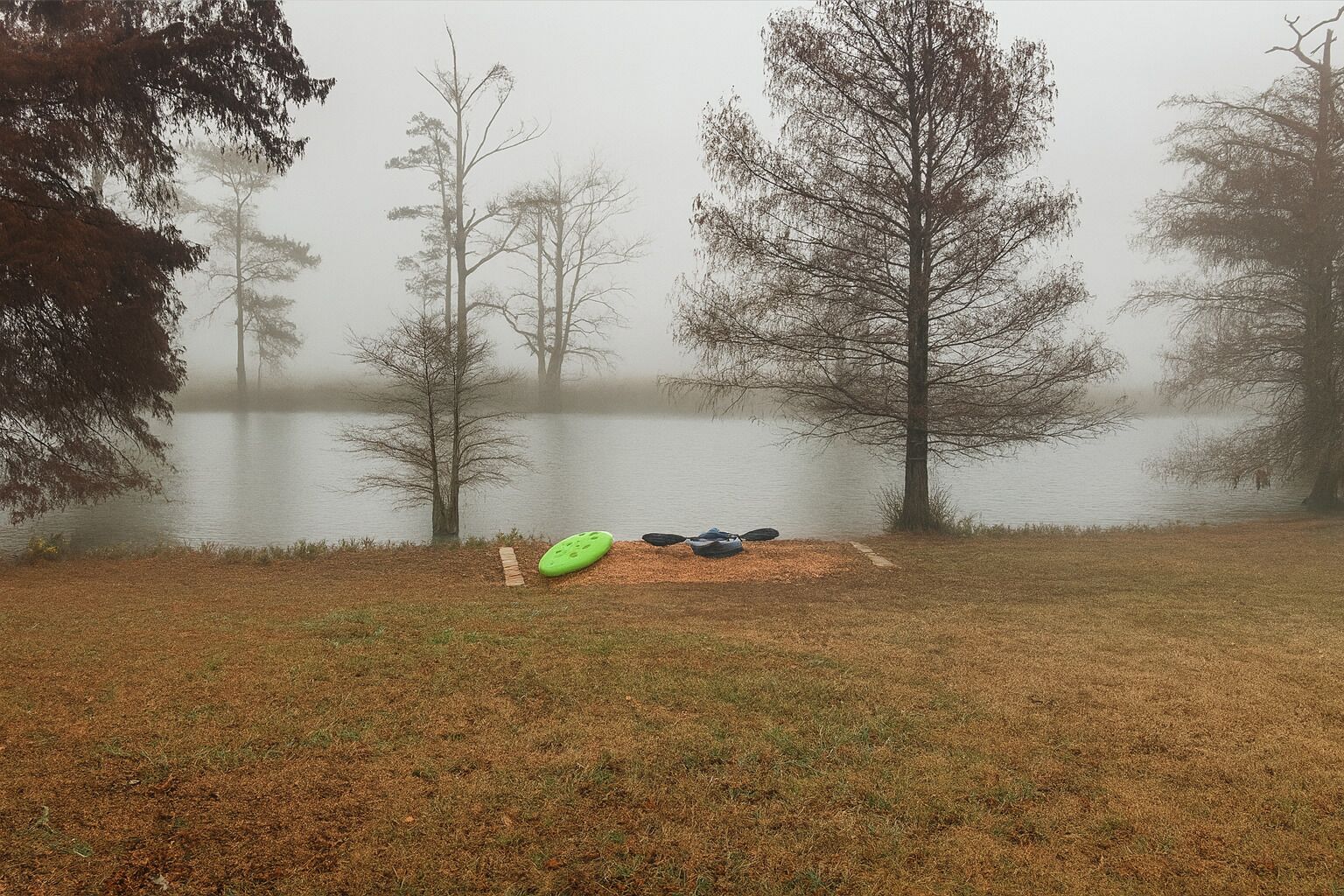 A foggy lake with trees and a grassy area with kayaks and a paddleboard.