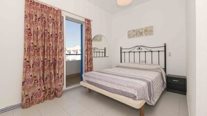 Un dormitorio con cama, ventana con cortinas, espejo y una pequeña estantería de pared.