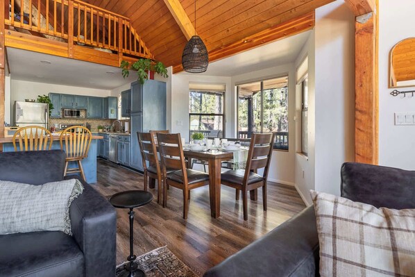 Un vaste espace de vie doté d’un plafond en bois, d’une table à manger pour quatre personnes et d’une cuisine aux meubles bleus.