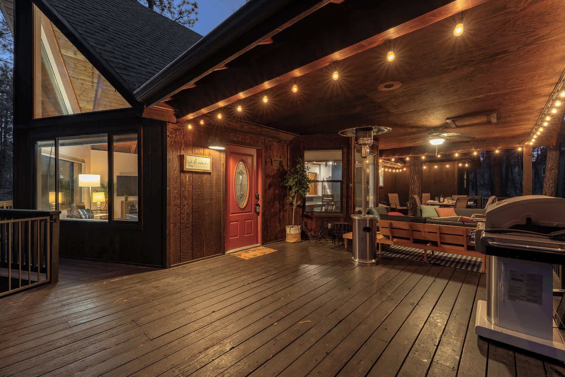 Una terraza de madera con una parrilla, zona de estar, una puerta roja y luces colgantes.