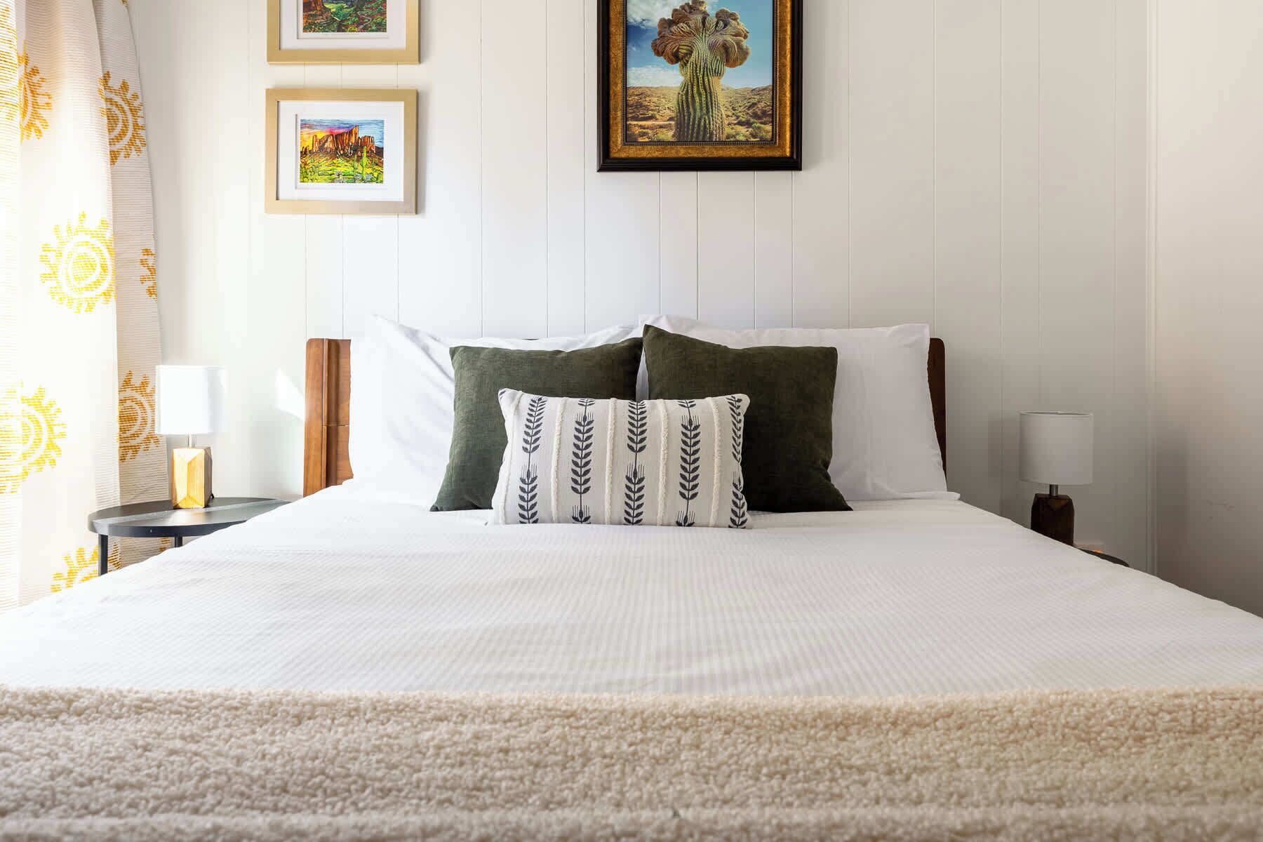 A neatly made bed with white bedding and two pillows, one green and one with a leaf pattern, in a room with white walls and a window with curtains.