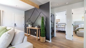 Ein modernes Wohnzimmer mit einer Couch, einem kleinen Holztisch mit Dekoration, einer hohen, grünen Pflanze und einer dunkelgrünen Wand mit geometrischen Mustern.