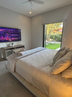 A bedroom with a large bed, a TV mounted on the wall, and a sliding glass door leading to a garden. - 3-bedroom house with private pool, AC, fitness center-5min from the sand. (Provincia de Guanacaste)