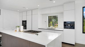 A modern kitchen with white cabinetry, a central island, and a window with a view of greenery. - Modern Luxury in Peaceful Hollywood Hills with Epic City Views (Los Angeles)