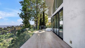 A modern balcony with a glass railing overlooking a lush landscape and cityscape. - Modern Luxury in Peaceful Hollywood Hills with Epic City Views (Los Angeles)