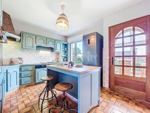 Eine Küche mit hellblauen Schränken, einer zentralen Kochinsel mit Hockern, einem großen Kühlschrank und einer Holztür mit Glasfeldern.