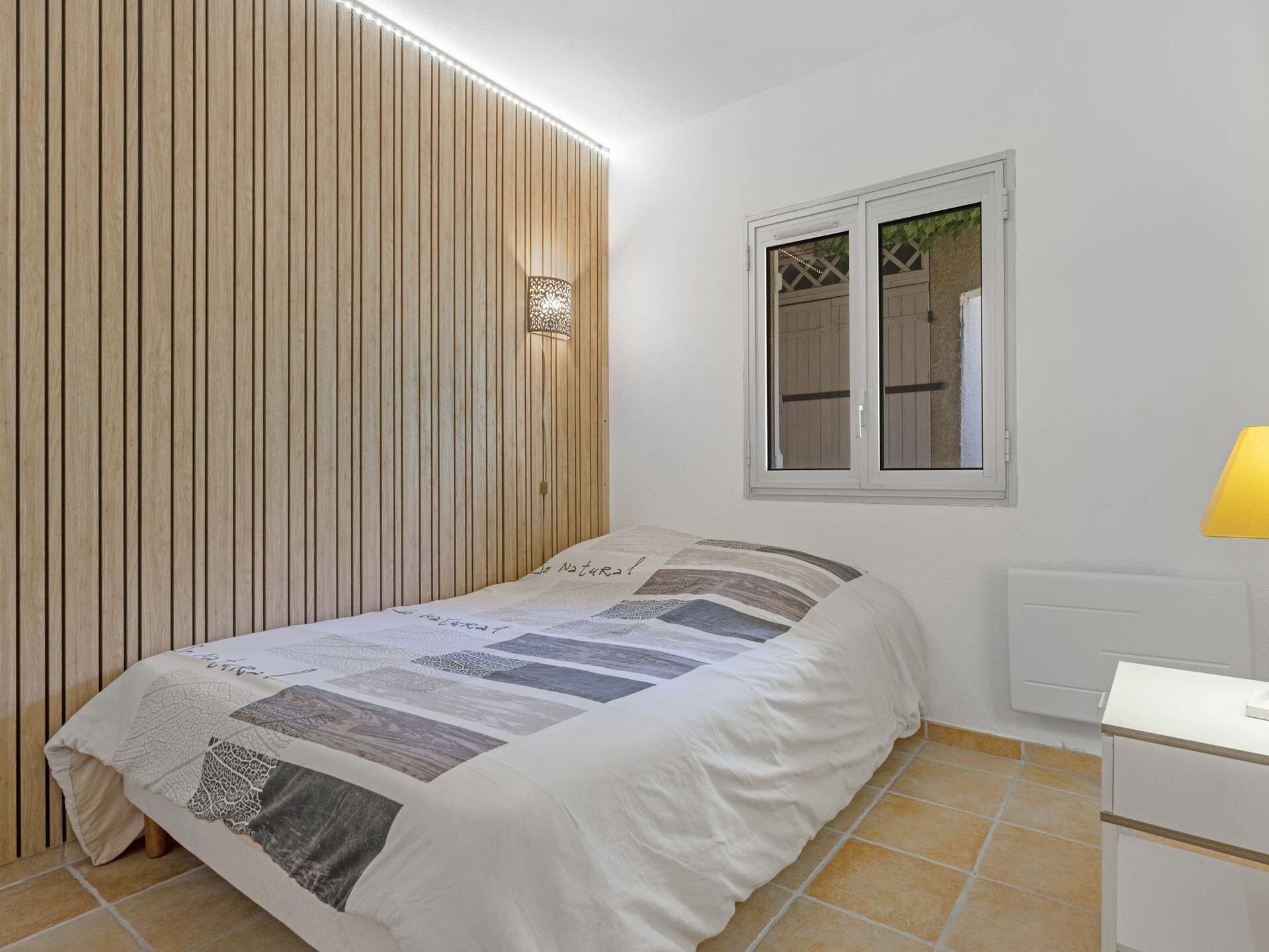 Un dormitorio con cama, ventana, luz empotrada en la pared y radiador.