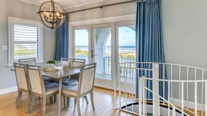 A dining area with a table and chairs, a white crib, and a view of the ocean through large windows. - Oceanfront Home! Spectacular Sunrise and Sunset Views, Private Boardwalk to Beach, Two Balconies! (St. Augustine)