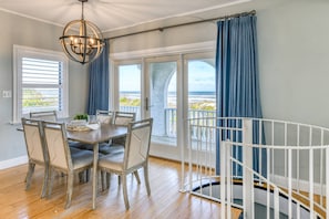 Ein Essbereich mit Tisch und Stühlen, einem weißen Babybett und Blick auf das Meer durch große Fenster.