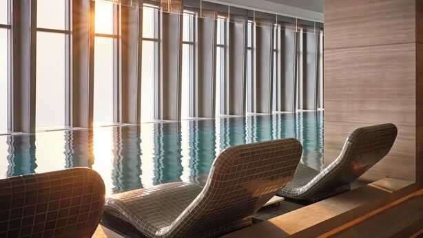 Une piscine intérieure moderne avec des murs en verre transparent, offrant une vue sur l’extérieur. On y trouve deux chaises longues à motifs à carreaux.