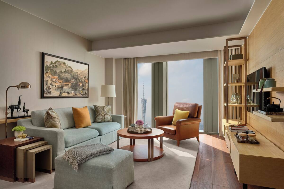 Ein modernes Hotelzimmer mit einer Sitzecke, bestehend aus Sofa, Sessel und Hocker, einem Couchtisch, einem Beistelltisch mit Lampe, einem Bücherregal und einem großen Fenster mit Blick auf die Stadt.