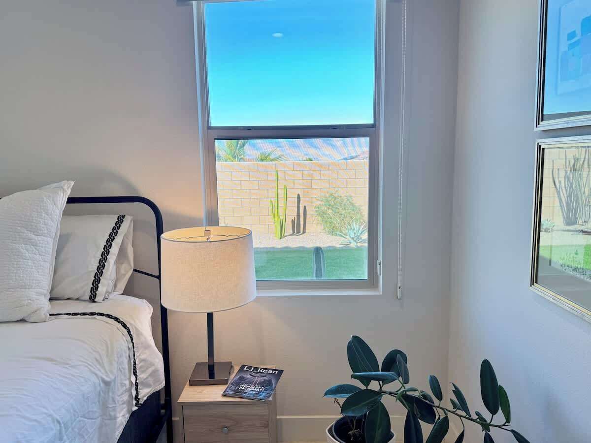 Ein Schlafzimmer mit einem Bett, einem Nachttisch mit Lampe, einem Fenster mit Blick auf einen Kakteengarten und einem gerahmten Bild an der Wand.