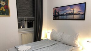 Una cama bien hecha con sábanas y toallas blancas, una mesita de noche con una lámpara, un cuadro de un paisaje urbano y una ventana con cortinas.