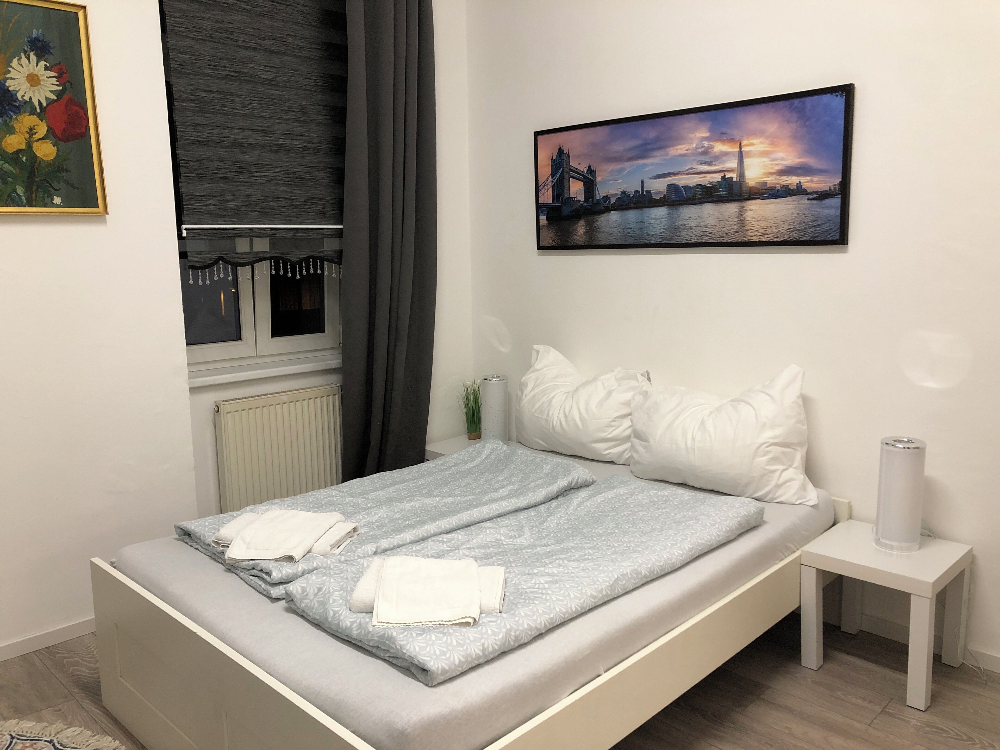 A neatly made bed with a grey and white patterned bedspread, white pillows, and folded towels on a white headboard. A framed cityscape painting hangs above the bed, and a small white nightstand with a lamp is to the right.